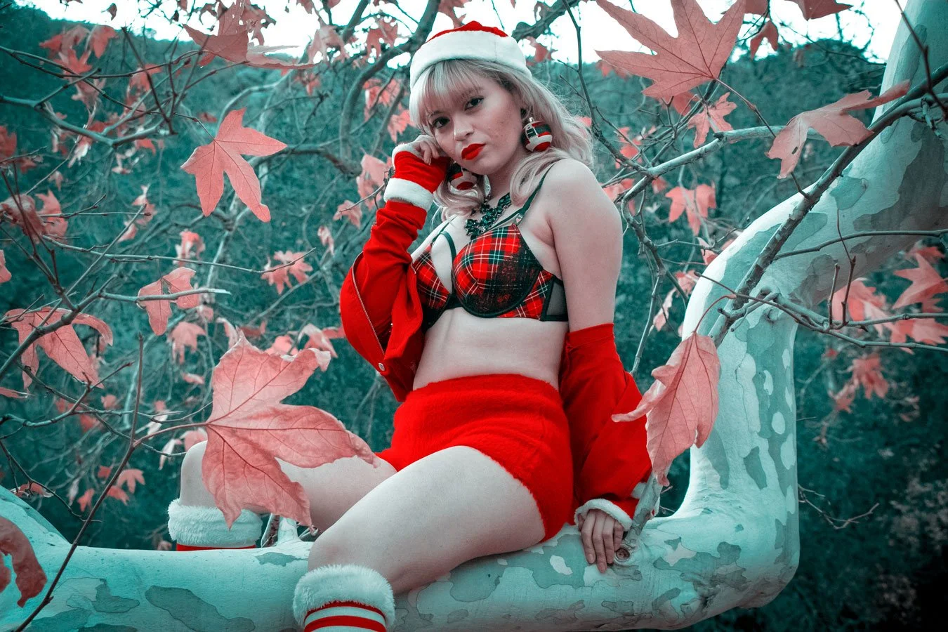 A woman in festive holiday attire, wearing a Santa hat, red gloves, red shorts, a plaid bra, and holiday accessories, sitting on a white tree swing surrounded by red autumn leaves.