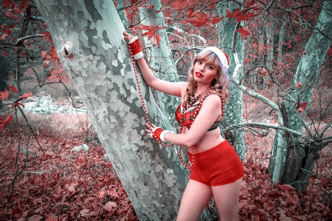 Woman dressed in a festive holiday outfit with a Santa hat, plaid top, red shorts, and holiday accessories poses by a tree in a forest with red leaves.