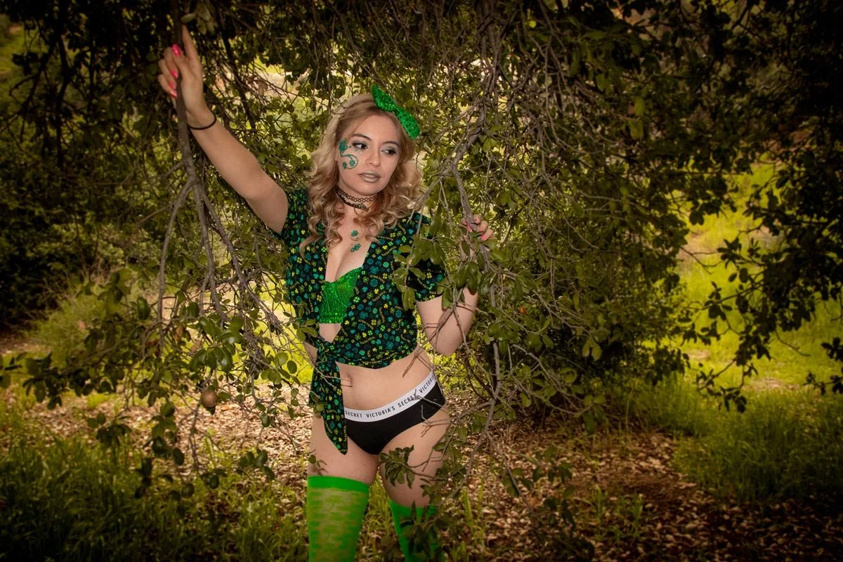 A woman with blonde curly hair, makeup, and tattoos posing outdoors among tree branches, wearing green lingerie, black Victoria's Secret underwear, a black and green floral top, green thigh-high stockings, and accessories like a green bow and choker.