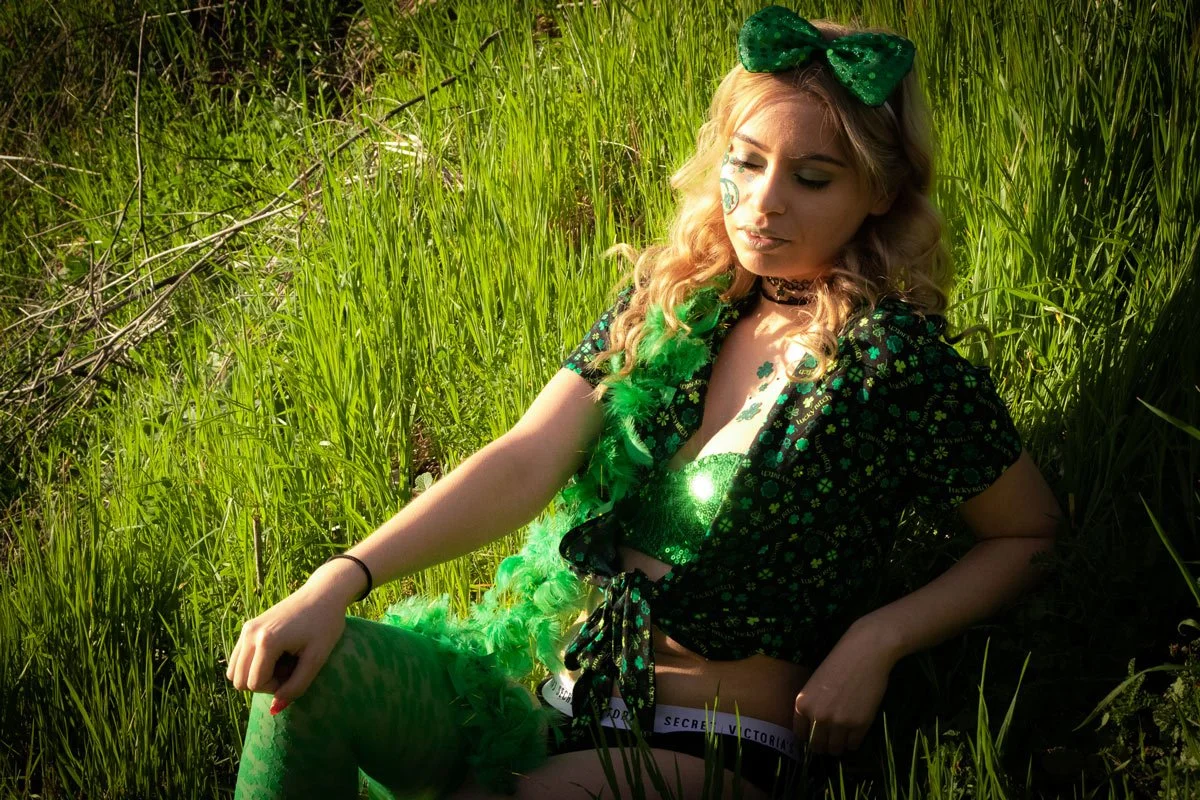 Young woman with blonde, wavy hair and green accessories, sitting in tall grass on a sunny day, wearing a black and green outfit.