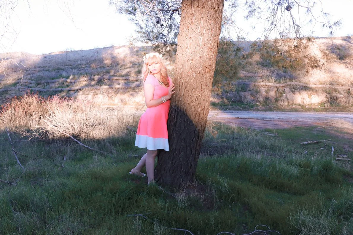 A woman with blonde hair and a pink dress standing outdoors near a tree in a grassy area with hills in the background.