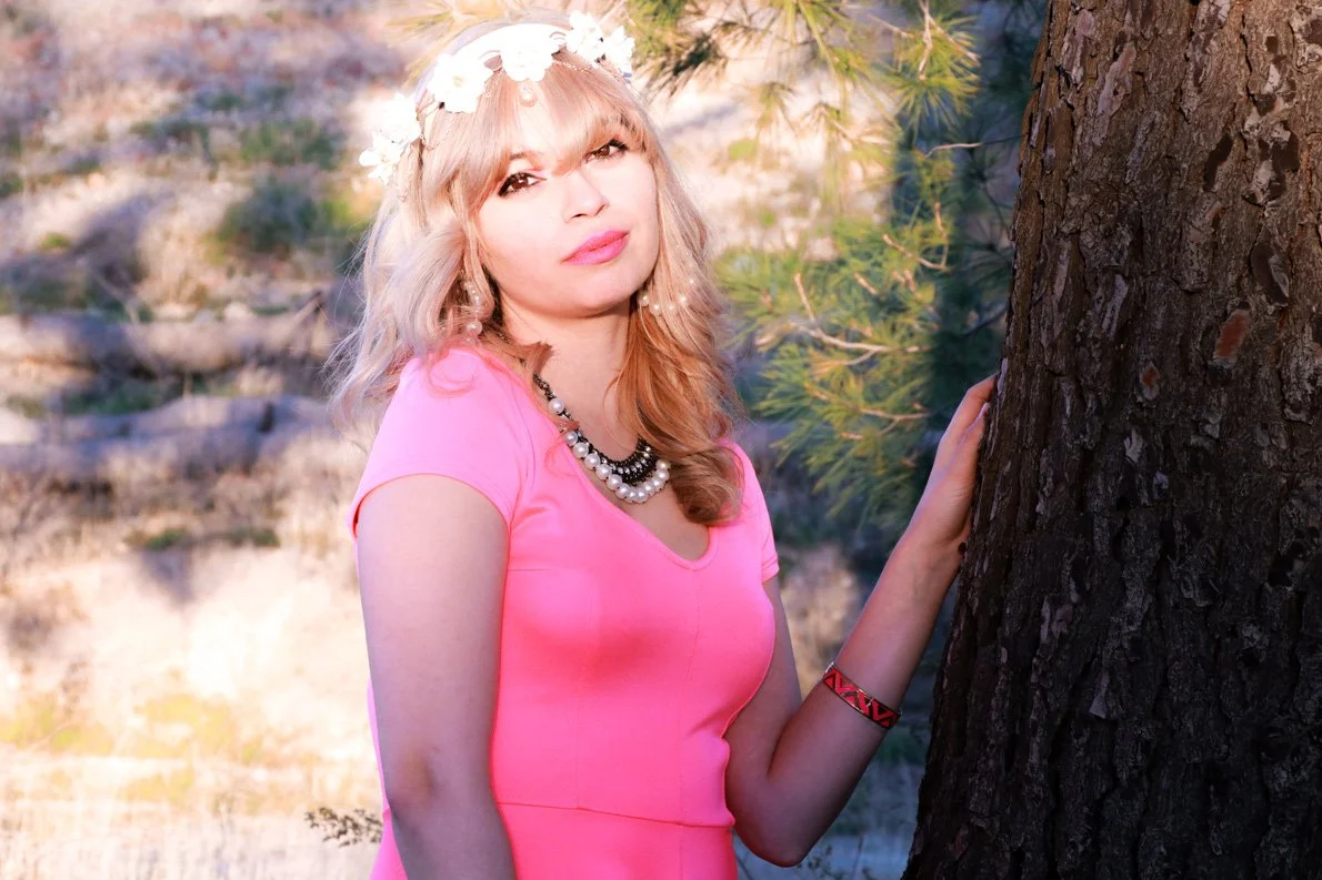 A woman with blonde hair, wearing a pink dress, a floral headband, and a pearl necklace, standing outdoors next to a tree and holding onto it.
