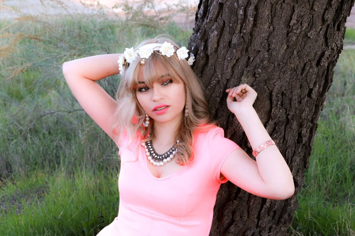 A young woman with blonde hair wearing a pink dress, floral headband, pearl necklace, earrings, and bracelet, posing against a tree in a grassy field.
