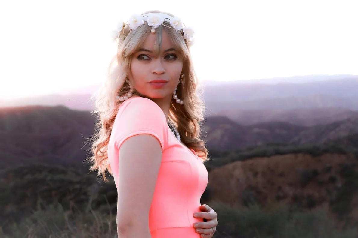 A young woman with blonde hair wearing a pink top, a white flower crown, and pearl earrings stands in front of a landscape with rolling hills during sunset.