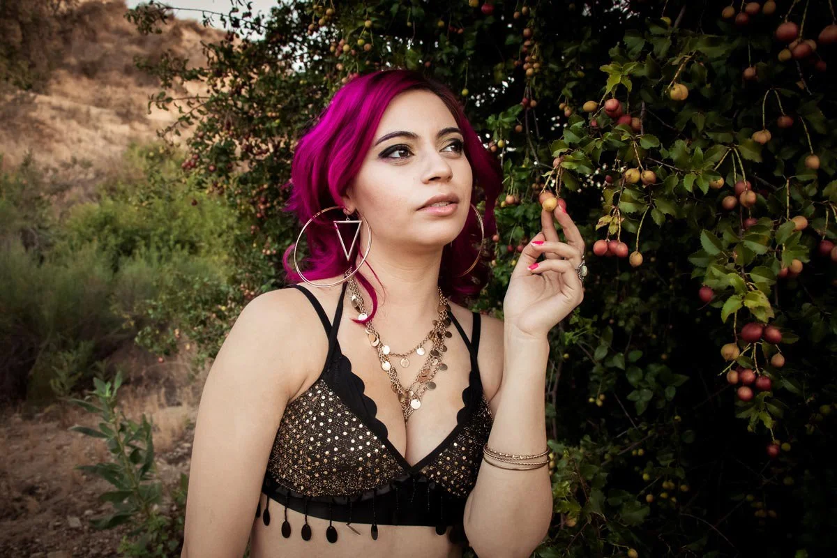 A woman with pink hair, wearing large hoop earrings and layered jewelry, standing in front of a bush with small round berries, holding a branch with berries in her hand.