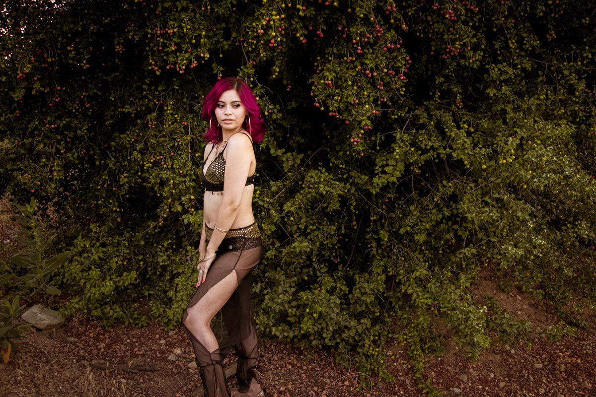 A young woman with magenta hair wearing black lingerie and sheer black pants stands outdoors in front of a dense bush with small red berries.
