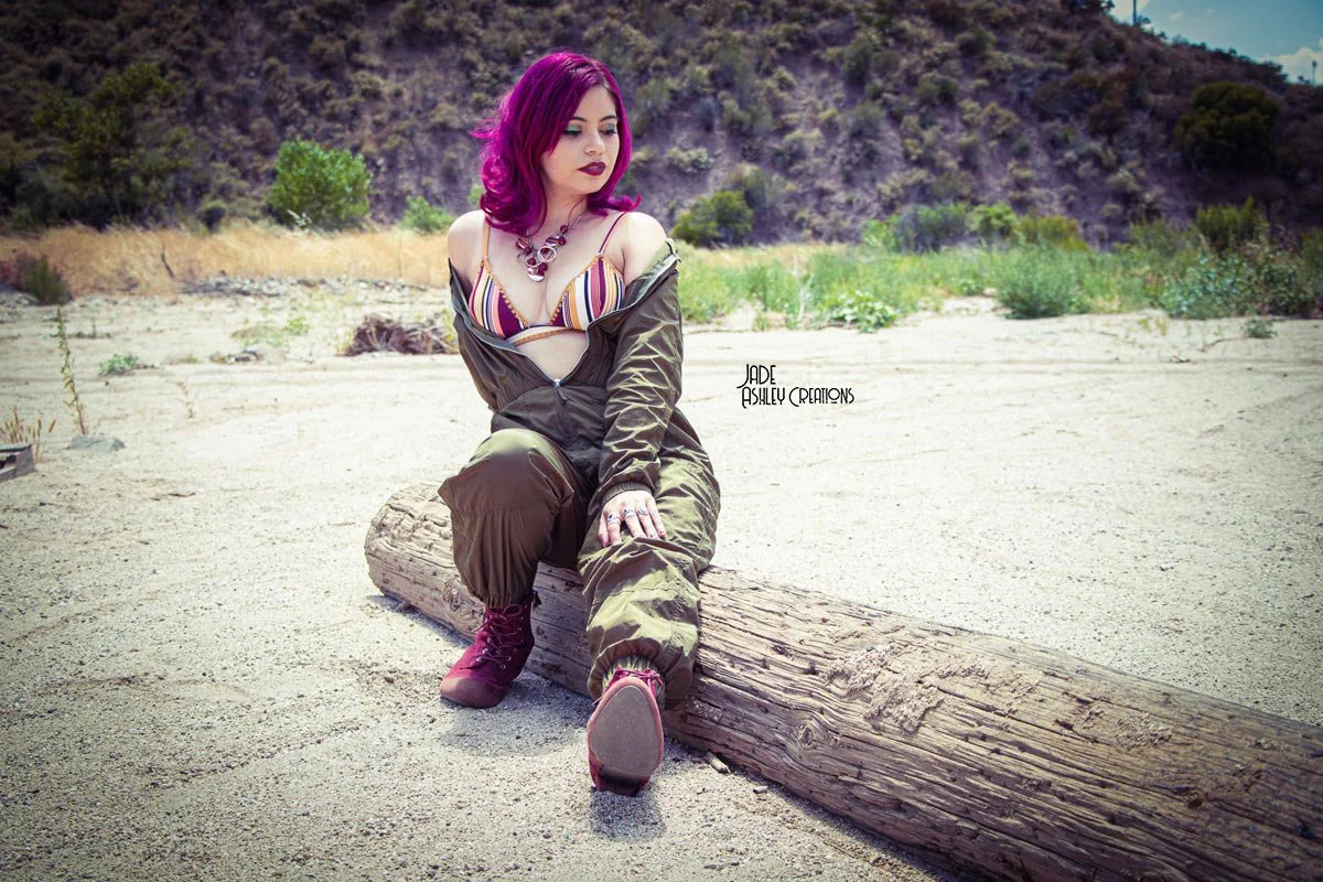 A woman with bright pink hair and a striped top sitting on a large log in a sandy, desert-like area with hills and sparse vegetation in the background.