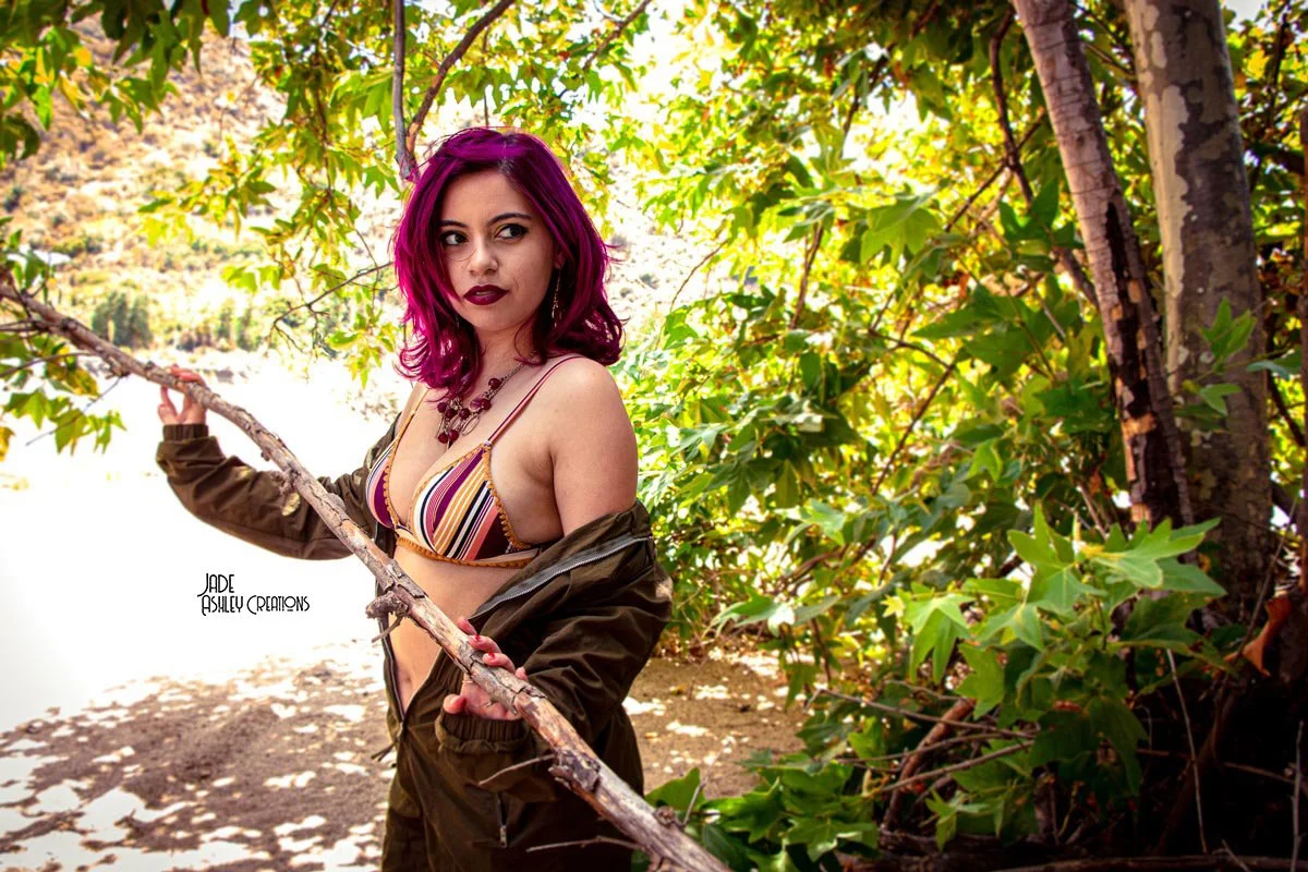 A woman with purple hair stands outdoors among greenery, wearing a striped bikini top and a partially unzipped jacket.
