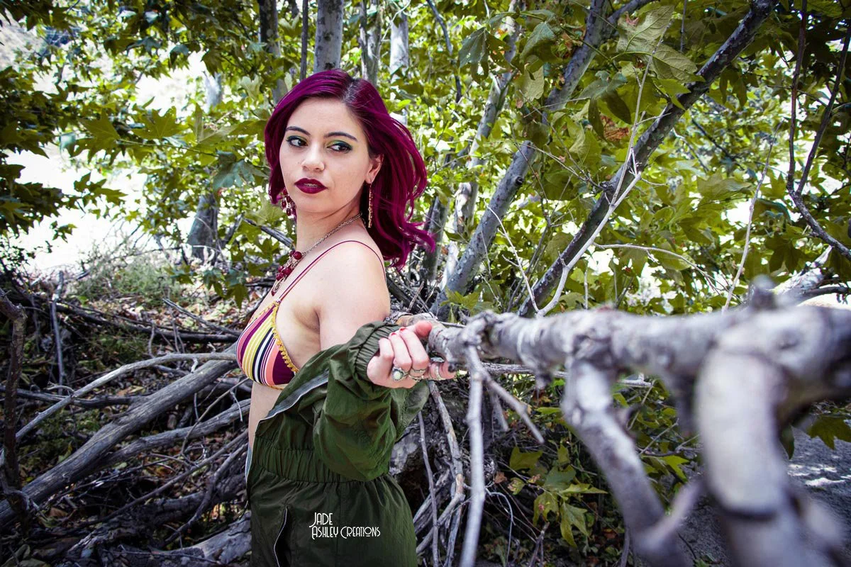 Young woman with dark pink hair and dark lipstick standing among tree branches and green leaves, wearing a colorful striped top, green jacket, and pants, looking at the camera.