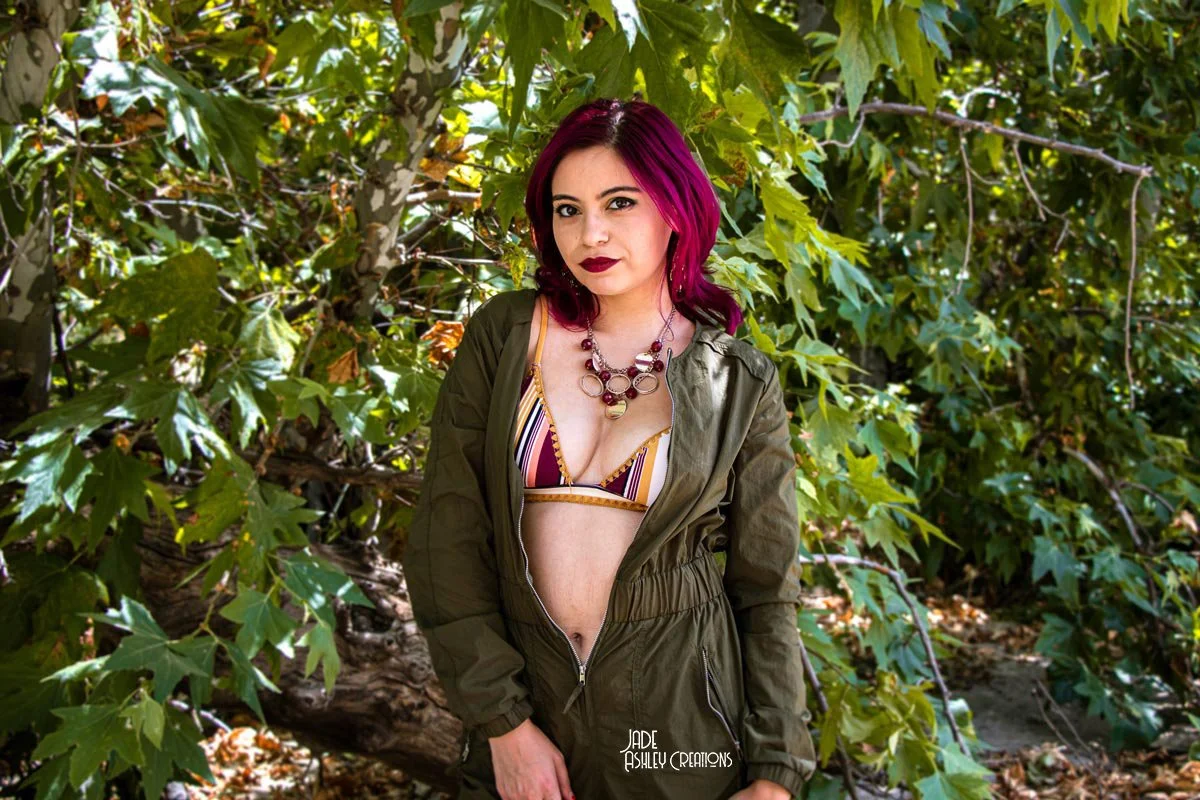 A woman with pink hair standing outdoors in front of green leafy trees, wearing a striped bikini top and an olive green jacket and pants, with a necklace, looking at the camera.
