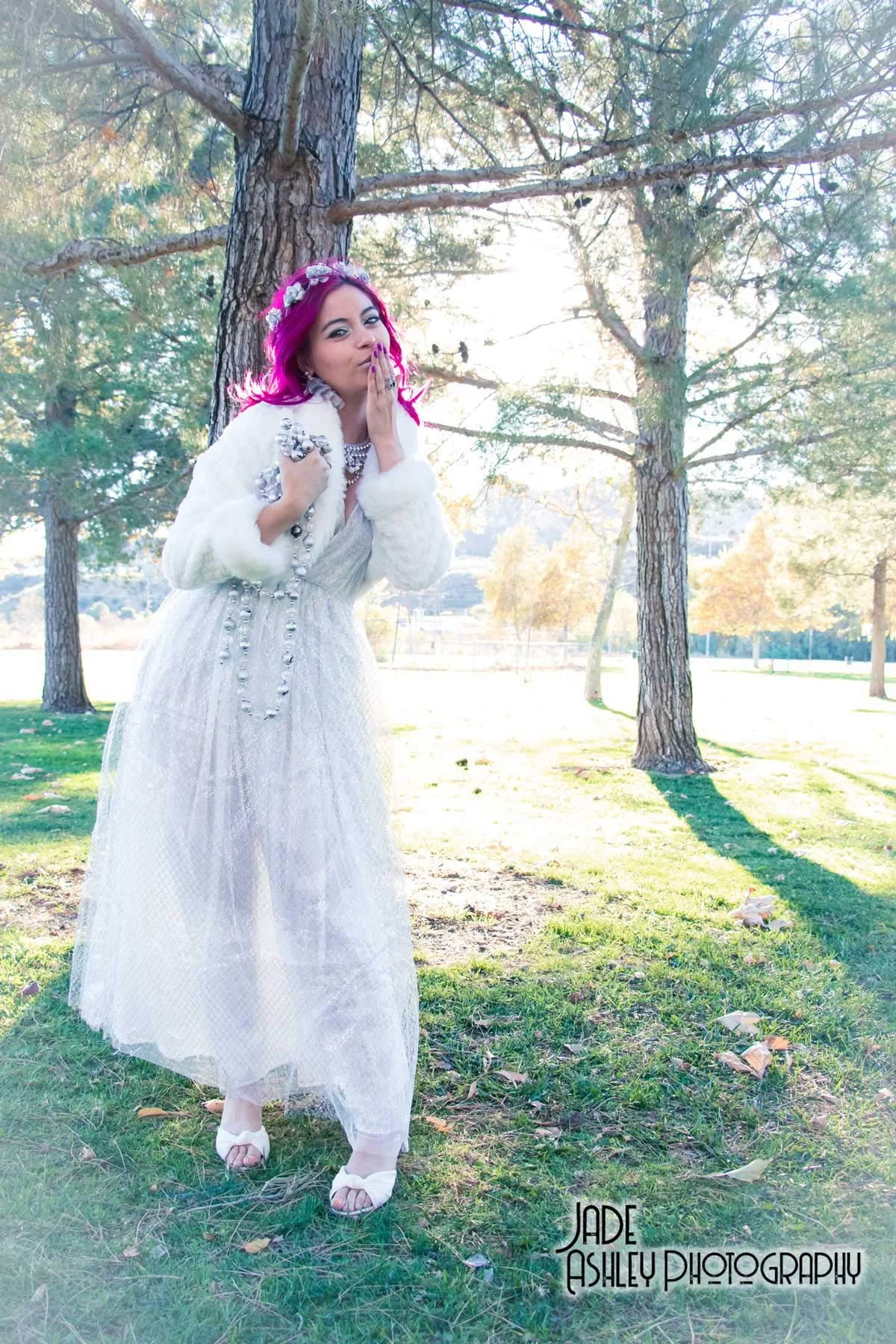 A woman with bright pink hair dressed in a white gown and floral crown, standing outdoors under trees with sunlight behind her, blowing a kiss.
