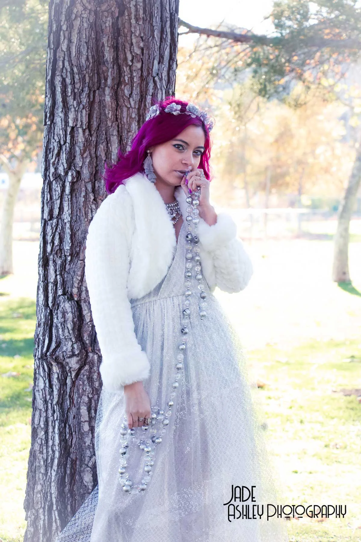 A woman with magenta hair standing outdoors next to a tree, holding a pearl necklace, wearing a white fur jacket over a silver dress with pearl embellishments, and a floral headband, in a park during sunny weather.