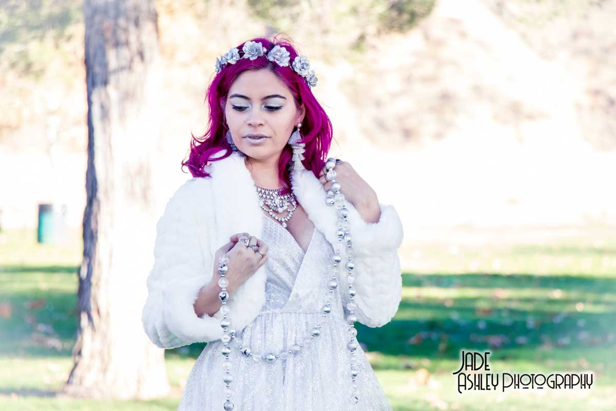 Woman with vibrant pink hair wearing a white dress, floral headband, and jewelry, standing outdoors holding a long string of pearls.