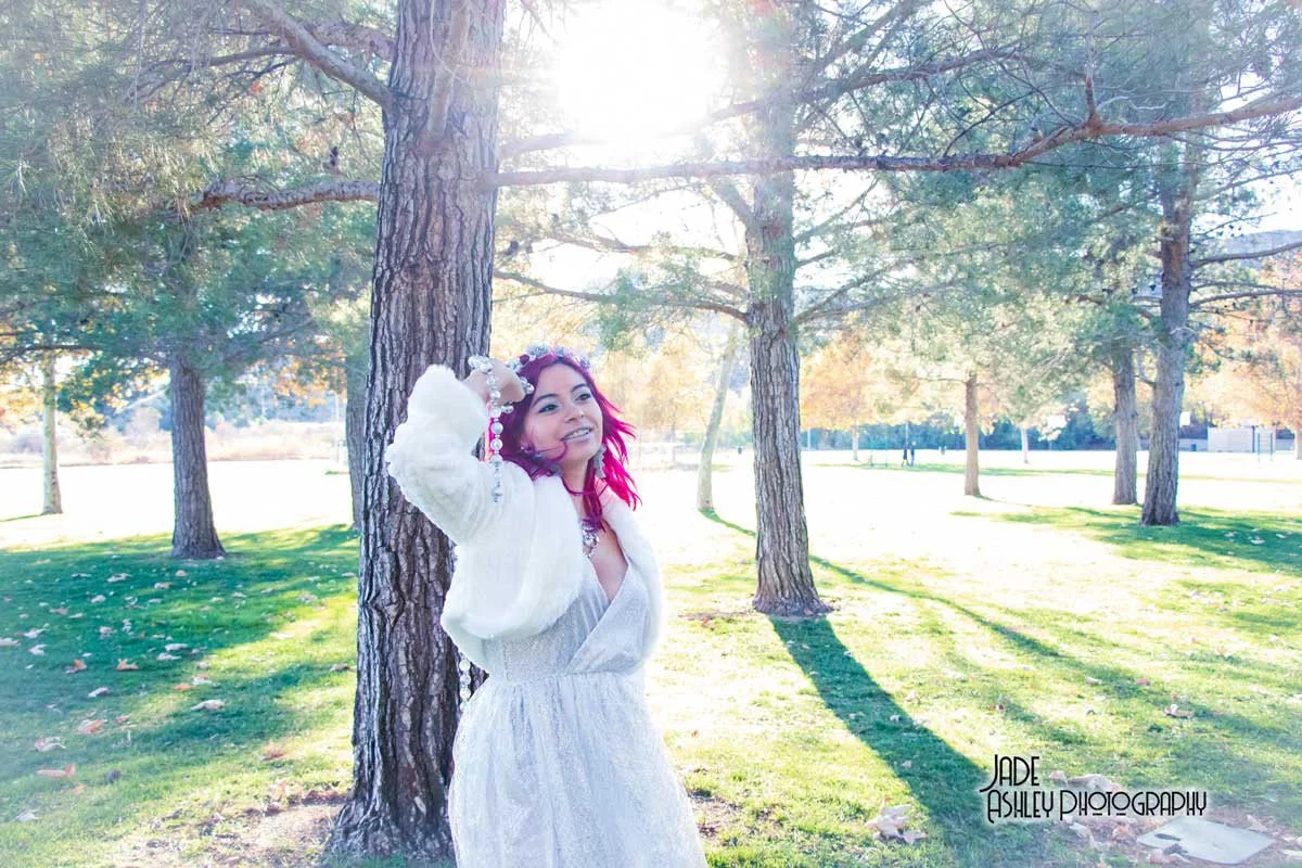 A woman with bright pink hair standing outdoors in a park with tall trees and lush green grass, smiling and posing with one hand on her head. The sunlight is shining brightly through the trees. The photo is marked with the text "Jade Ashley Photograp