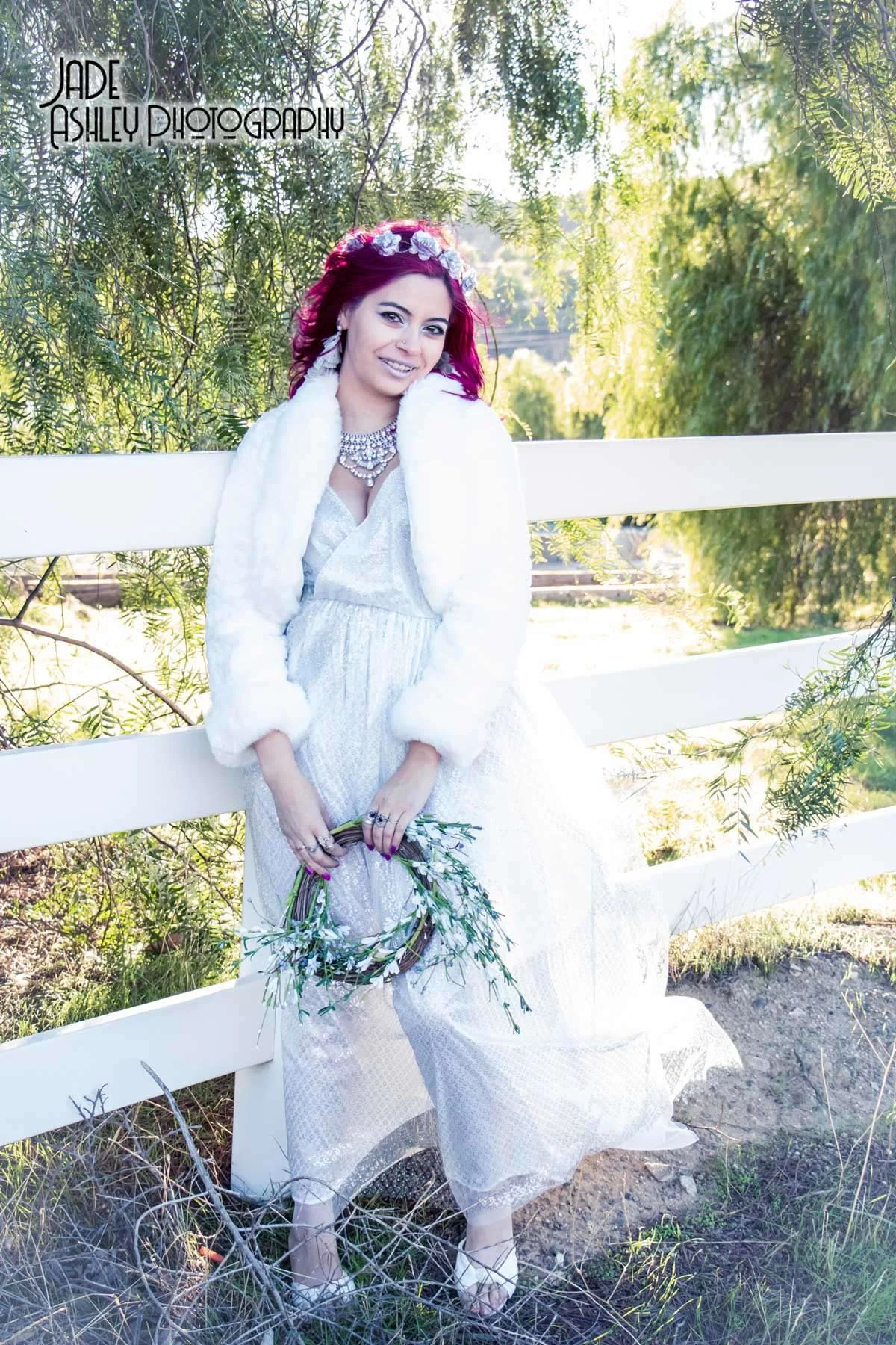A woman with vibrant red hair, gold hoop earrings, and a floral headband, dressed in a white dress with lace details, accessorized with a statement necklace, is standing outdoors beside a white fence, holding a small bouquet of greenery, with green t