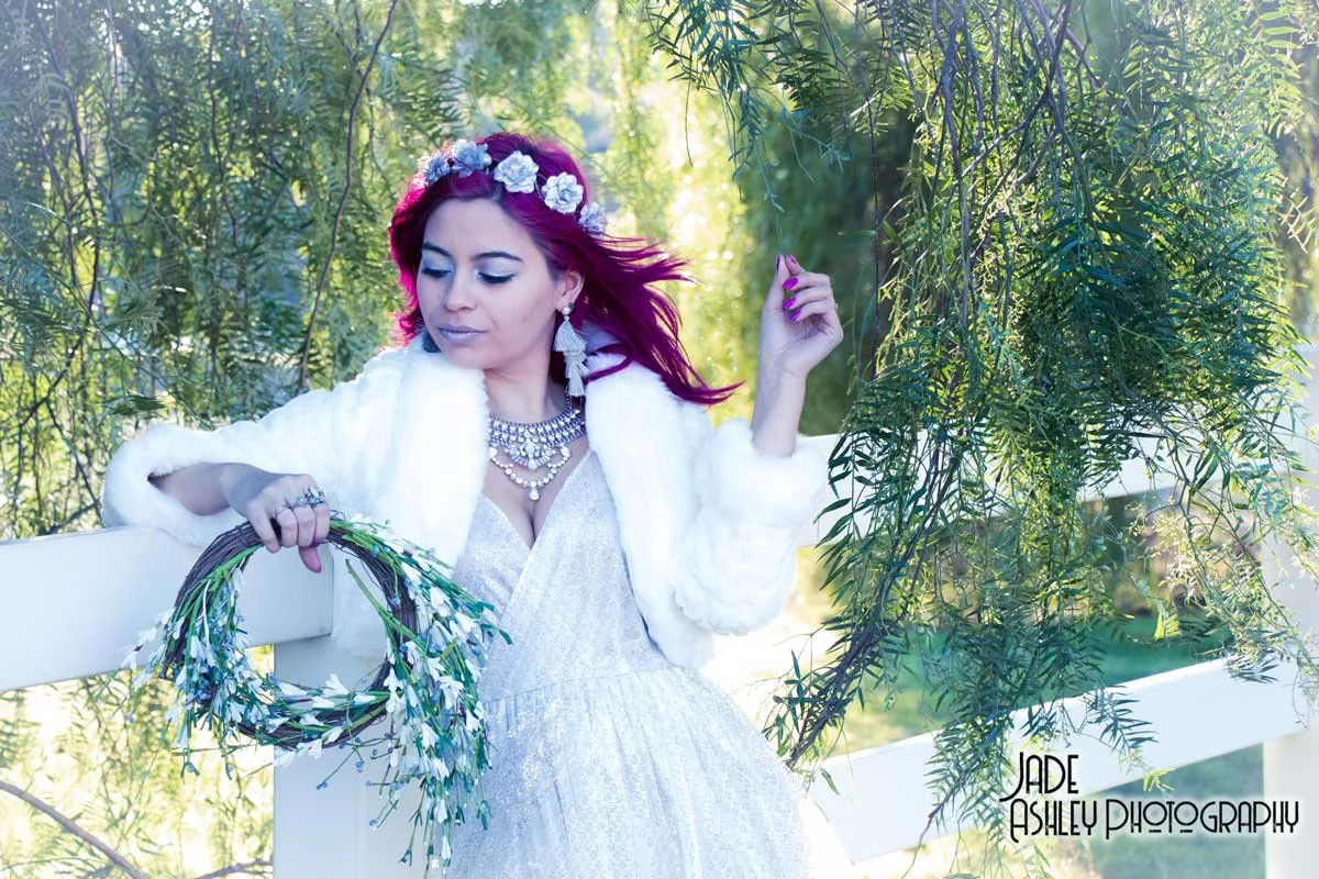 A woman with pink hair, wearing a white fur coat and a white dress, is outdoors surrounded by greenery. She is holding a wreath made of white and green flowers and wearing a floral headband and jewelry.