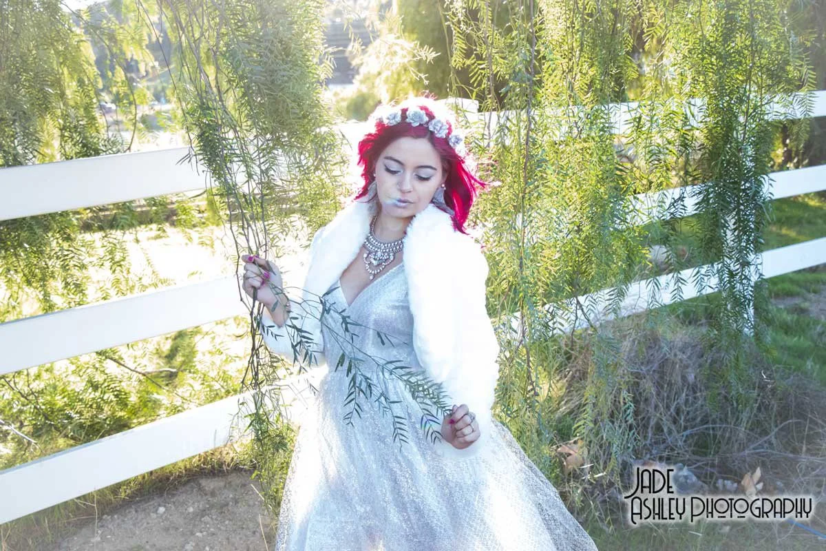 A woman with red hair wearing a silver gown, white fur stole, and floral crown standing outdoors near a white fence and green foliage, looking down peacefully.