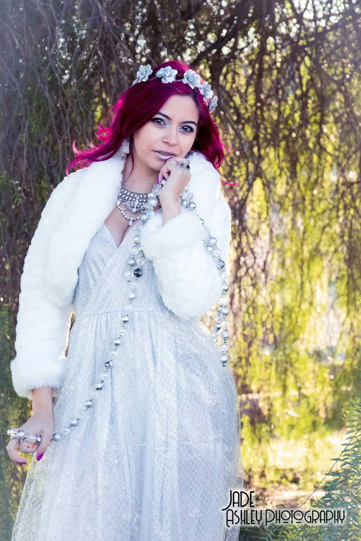 Young woman with vibrant purple hair and flowers in her hair, standing outdoors in a natural, wooded setting. She is dressed in a shiny silver gown, layered with a white faux fur jacket, and accessorized with large pearl jewelry and rings.
