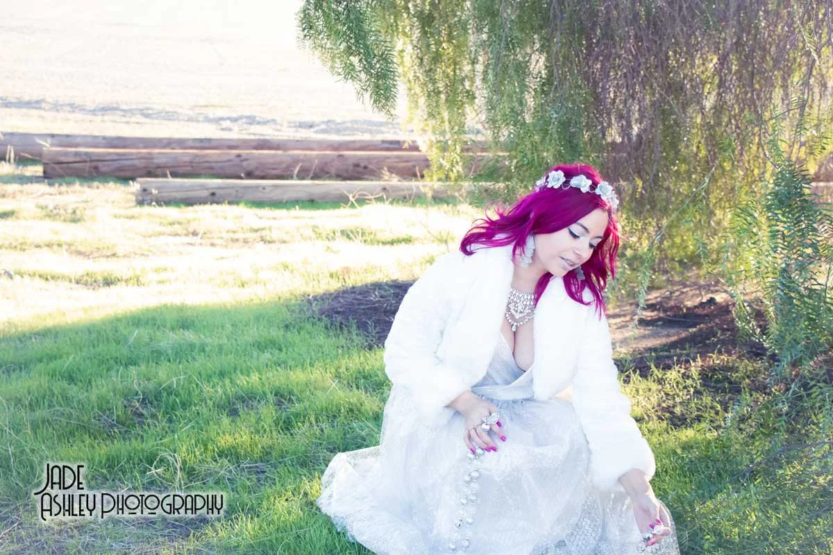 A woman with magenta hair sitting outdoors under a tree, wearing a white dress and jewelry, with a floral headband and a serene expression.
