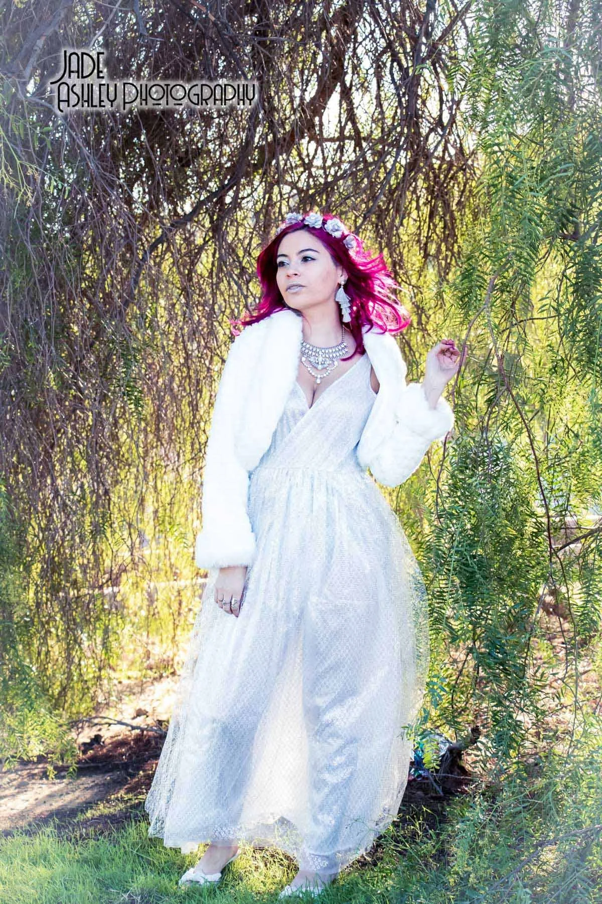 A woman with vibrant pink hair stands outdoors amidst greenery and tree branches, dressed in a white gown, with jewelry and a white fur coat, posing gracefully.