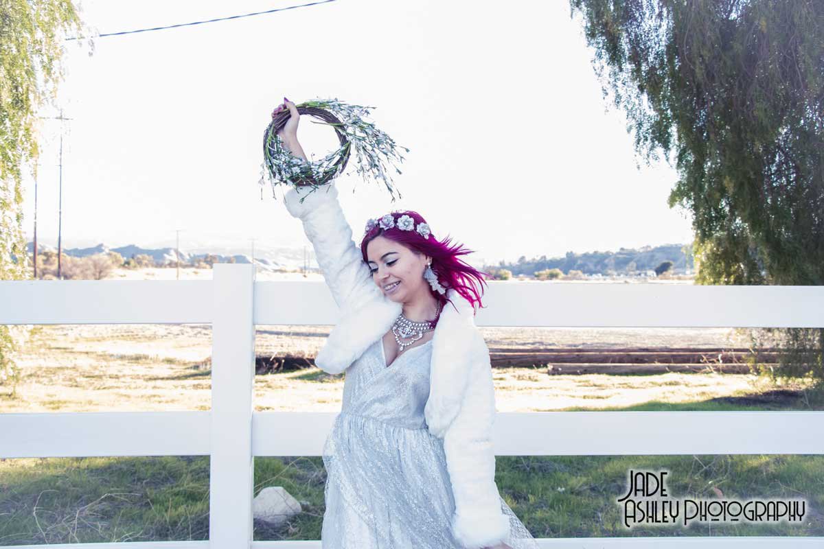 Woman with pink hair and floral headband smiling with eyes closed, holding a wreath, standing outdoors in front of a white fence.