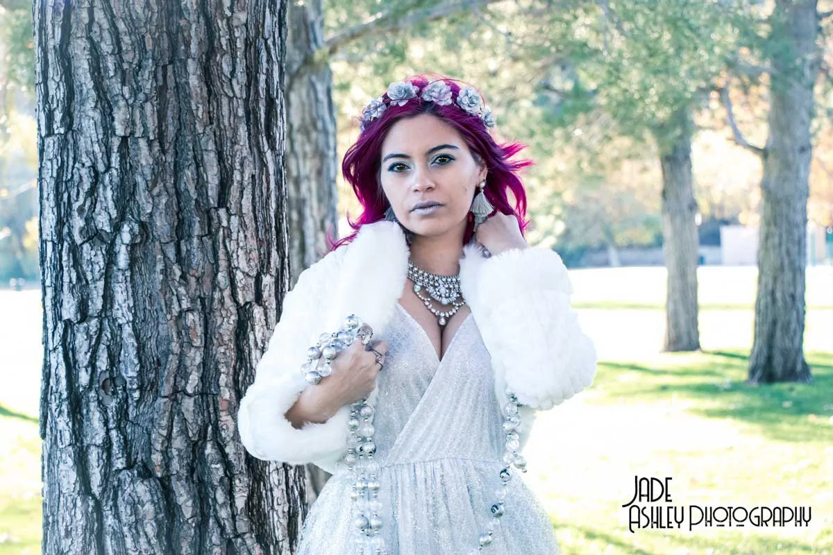 Woman with magenta hair and a flower crown, wearing a white dress and jewelry, standing outdoors next to a tree in a park.