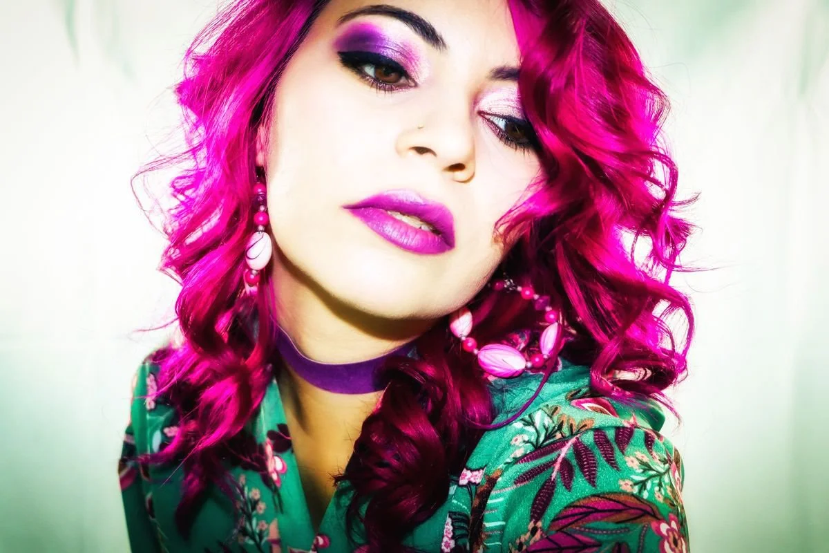 A woman with bright pink, curly hair and vibrant makeup, wearing pink and white jewelry, a green patterned shirt, and a purple choker.