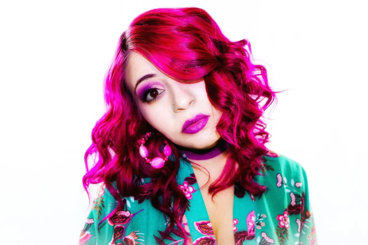 A young woman with vibrant pink hair styled in loose curls, wearing colorful makeup and bold purple lipstick, accessorized with large pink earrings and a floral green top, posing with her head tilted slightly, against a plain white background.