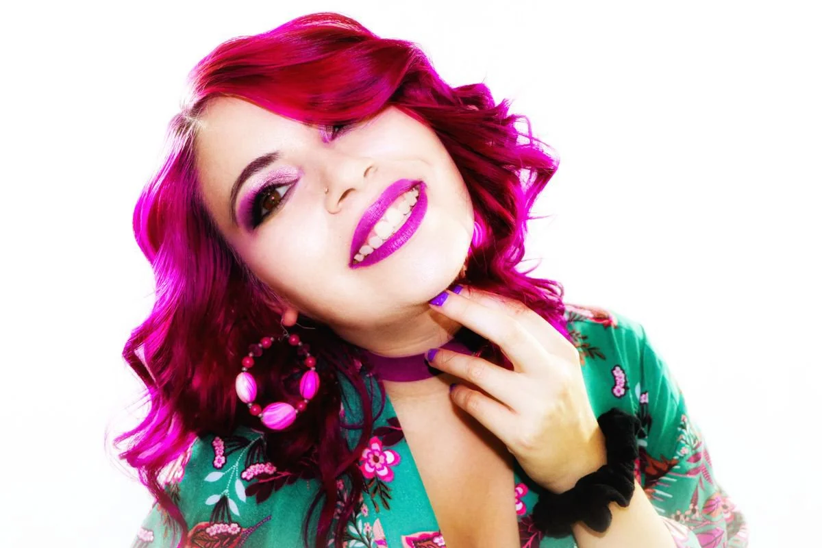A woman with bright pink hair styled in curls, wearing bold makeup with purple eyeshadow and matching lipstick. She's smiling revealing white teeth, with one eye partially closed, and has a nose piercing. She is dressed in a colorful floral top, larg