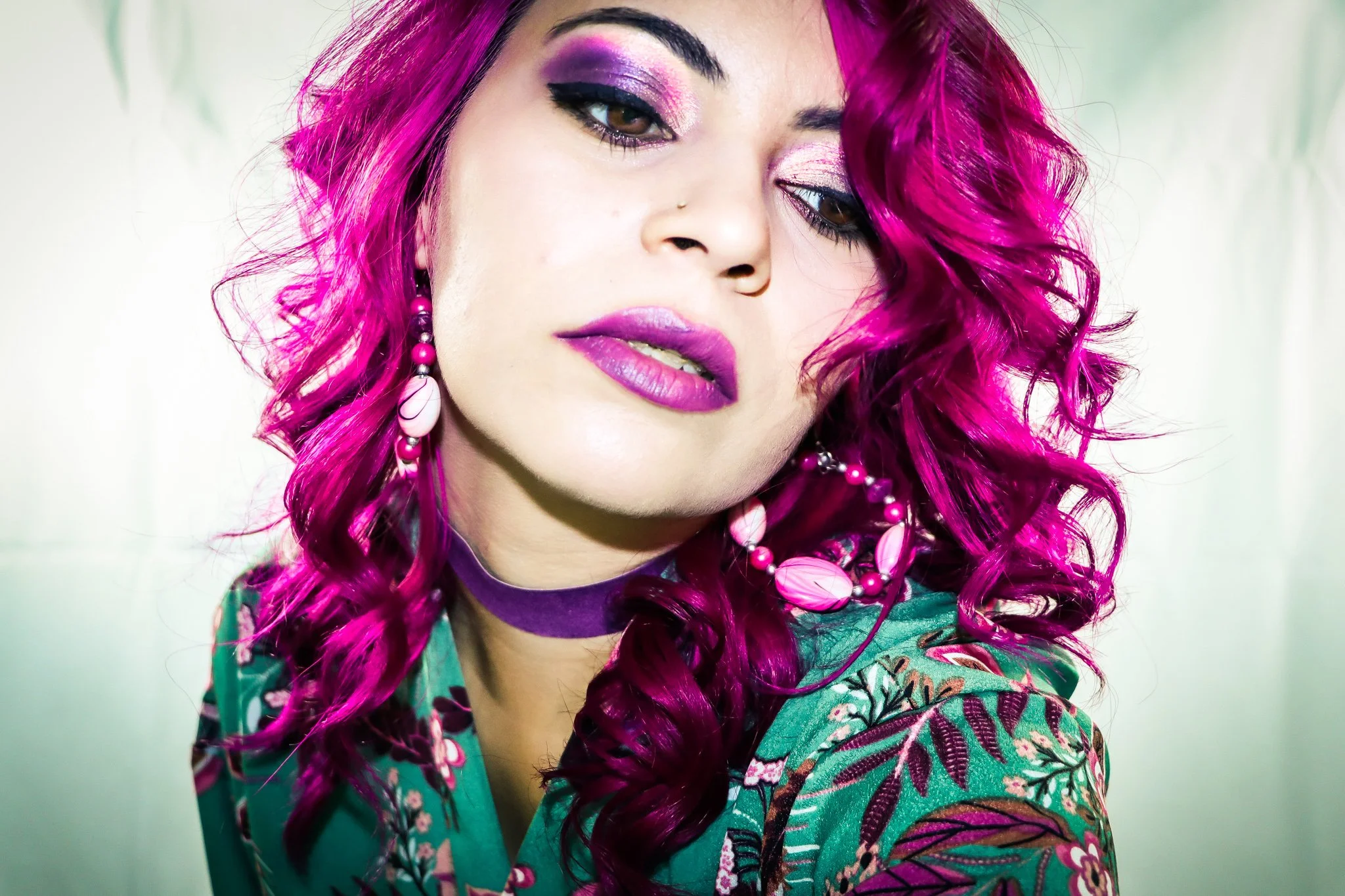 A woman with bright pink curly hair, pink lipstick, colorful eye makeup, and wearing pink earrings, a purple choker, and a floral green top.