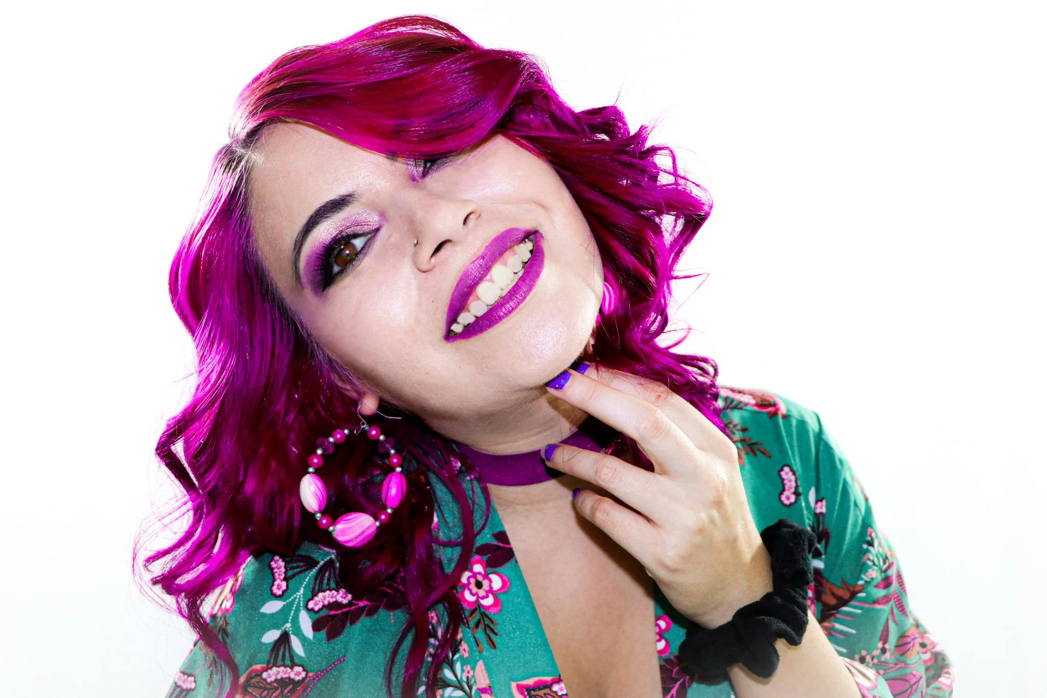 Woman with bright pink and purple hair, wearing purple lipstick, earrings, and a colorful floral top, smiling with one hand near her chin against a white background.