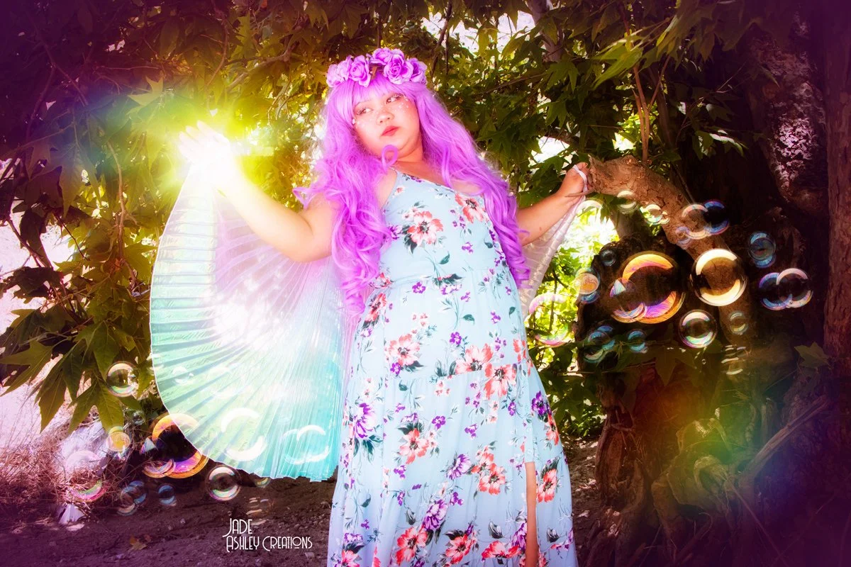 A young girl dressed as a fairy with pink hair, a flower crown, and wings, standing among trees with sunlight behind her and bubbles floating around.
