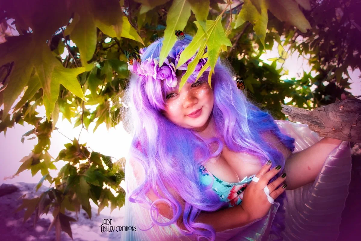 A woman with long, wavy purple hair and a flower crown, surrounded by green leaves and butterflies, smiling and posing outdoors.