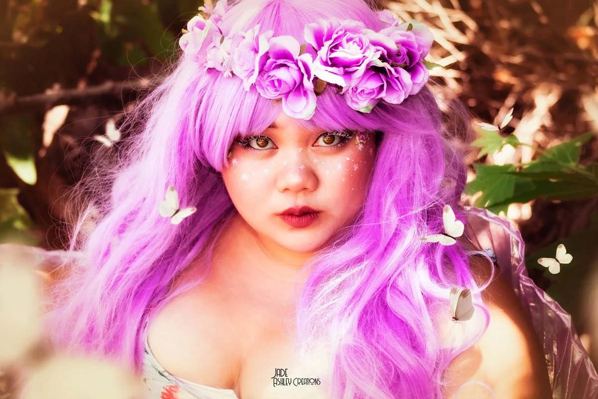 Young woman with long, vibrant purple hair and a floral crown made of purple roses, surrounded by butterflies, in an outdoor setting.
