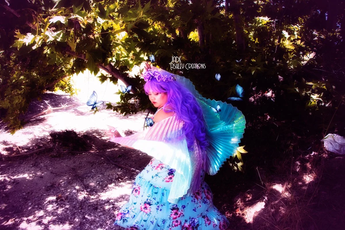 A young woman with long purple hair, wearing a flower crown and light-colored striped blouse, sitting amidst green foliage and butterflies, holding a large, blue, iridescent fan.