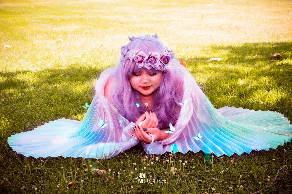 A girl with lavender hair wearing a flower crown, lying on the grass with her hands together, surrounded by glowing blue butterflies and wearing a flowing, iridescent dress.