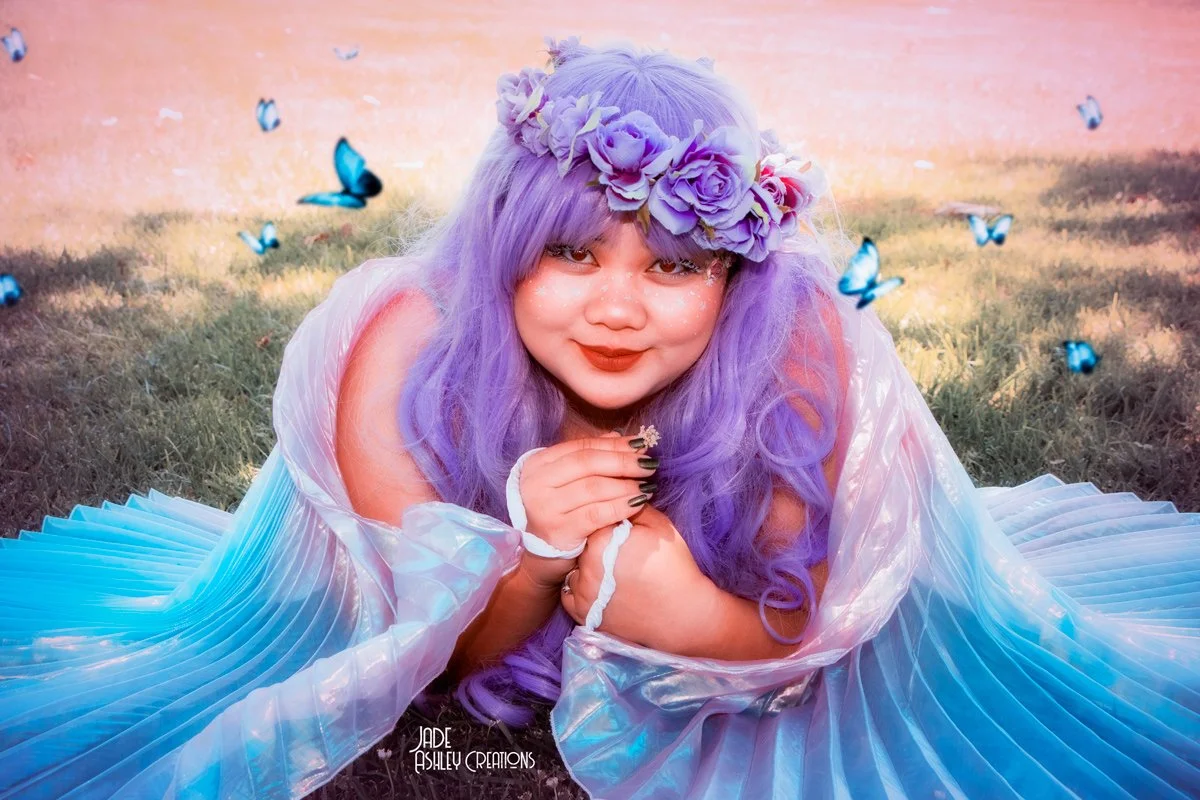 Young woman with purple hair and flower crown, wearing a flowing iridescent dress, lying on grass with butterflies flying around in a dreamy outdoor setting.