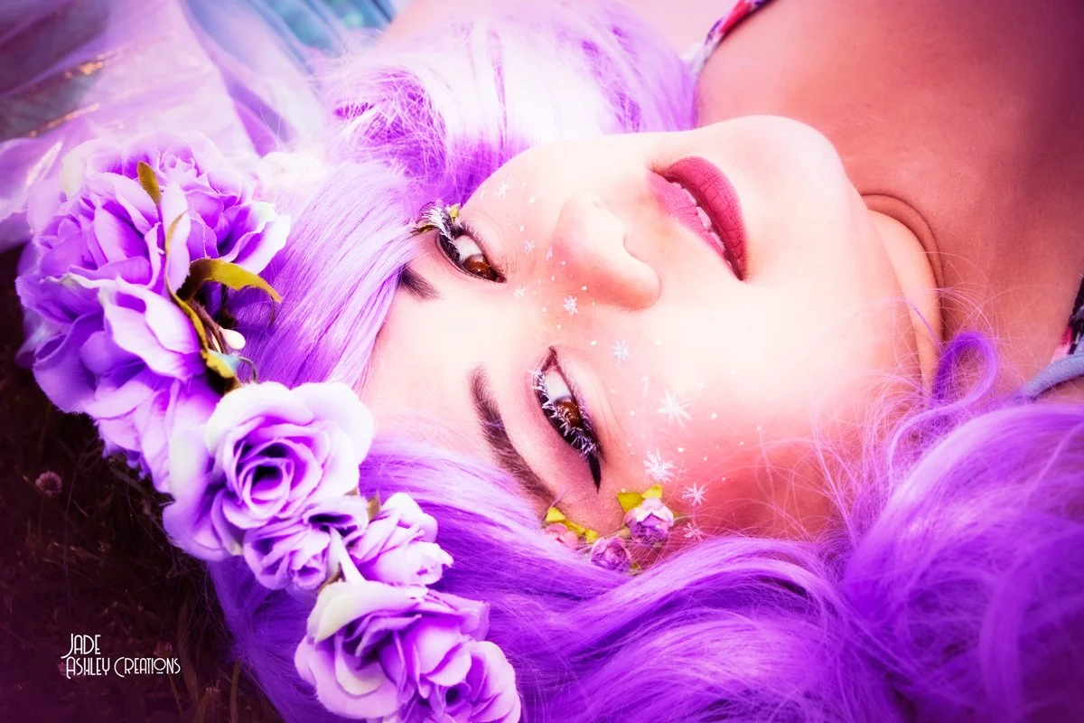 A young woman with long, wavy purple hair lying on her back, wearing a flower crown made of purple roses and other flowers, with her eyes open and makeup featuring white snowflake designs around her eyes.