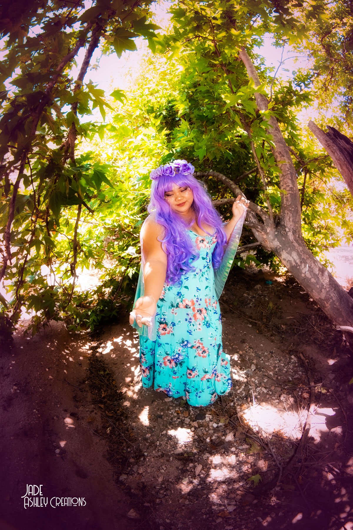 A woman with long purple hair and a flower crown, wearing a flowing, blue floral dress, standing outdoors under a leafy tree with sunlight filtering through the branches.