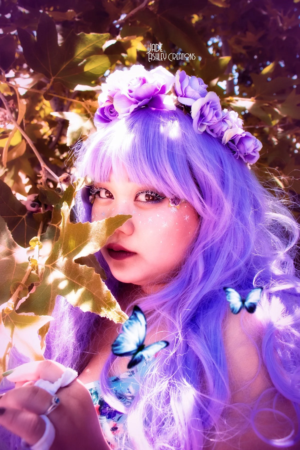 A woman with long, wavy purple hair wearing a flower crown made of purple flowers. She has makeup with sparkles on her cheeks and dark lipstick, and is surrounded by green leaves and blue butterflies.