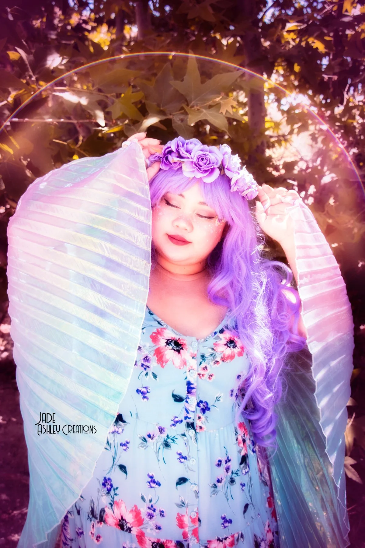 A woman with long purple hair wearing a floral dress, a purple flower crown, and holding an iridescent fan in front of leafy background