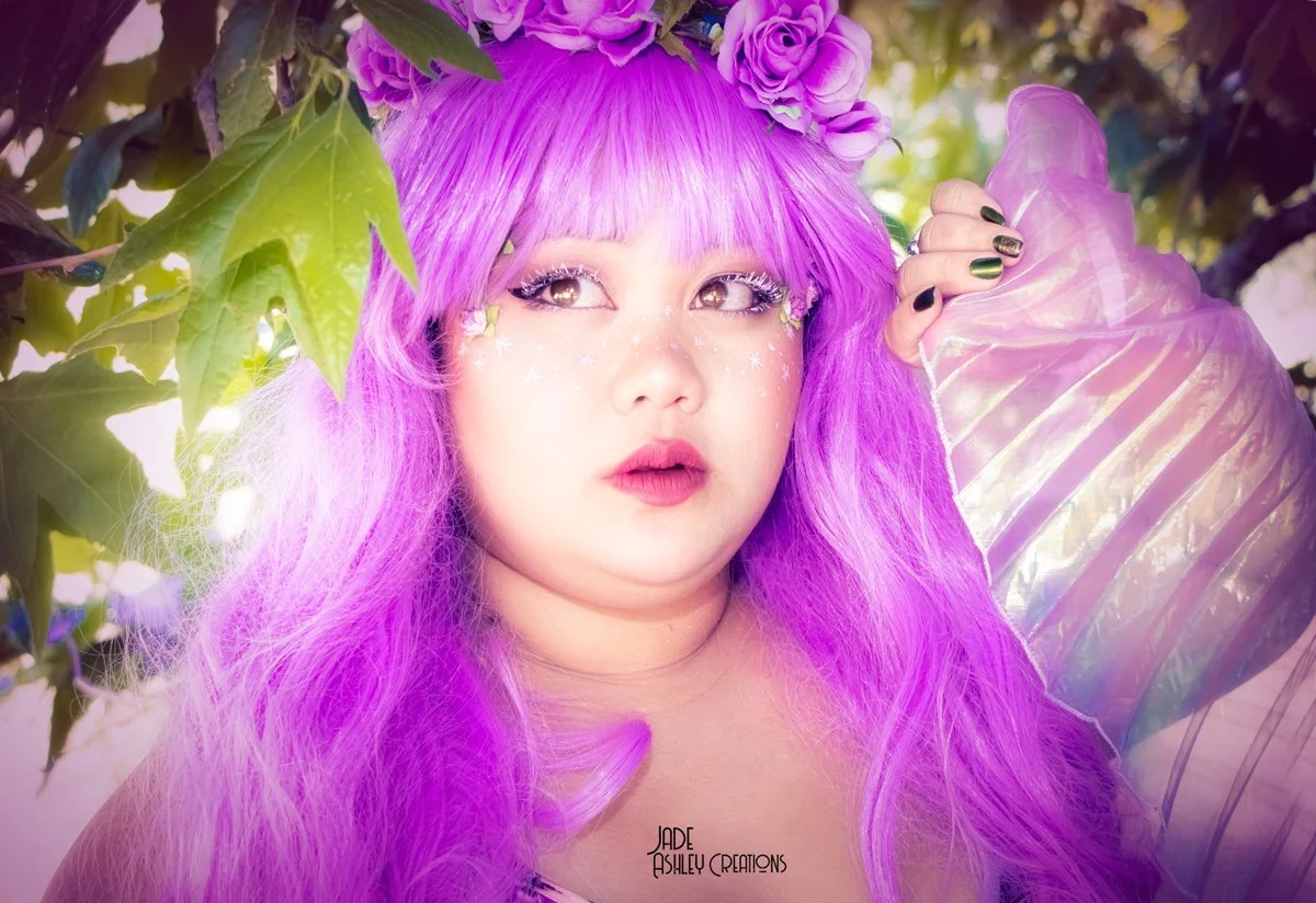 A woman with vibrant purple hair and flower crown posing among green leaves, holding a large iridescent seashell close to her ear, with glitter and star-shaped makeup accents around her eyes.