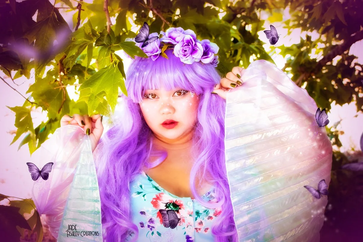 A young woman with long, wavy purple hair and a purple flower crown, standing among green leaves with butterflies around her. She has makeup with sparkles and is wearing a floral dress with iridescent butterfly wings.