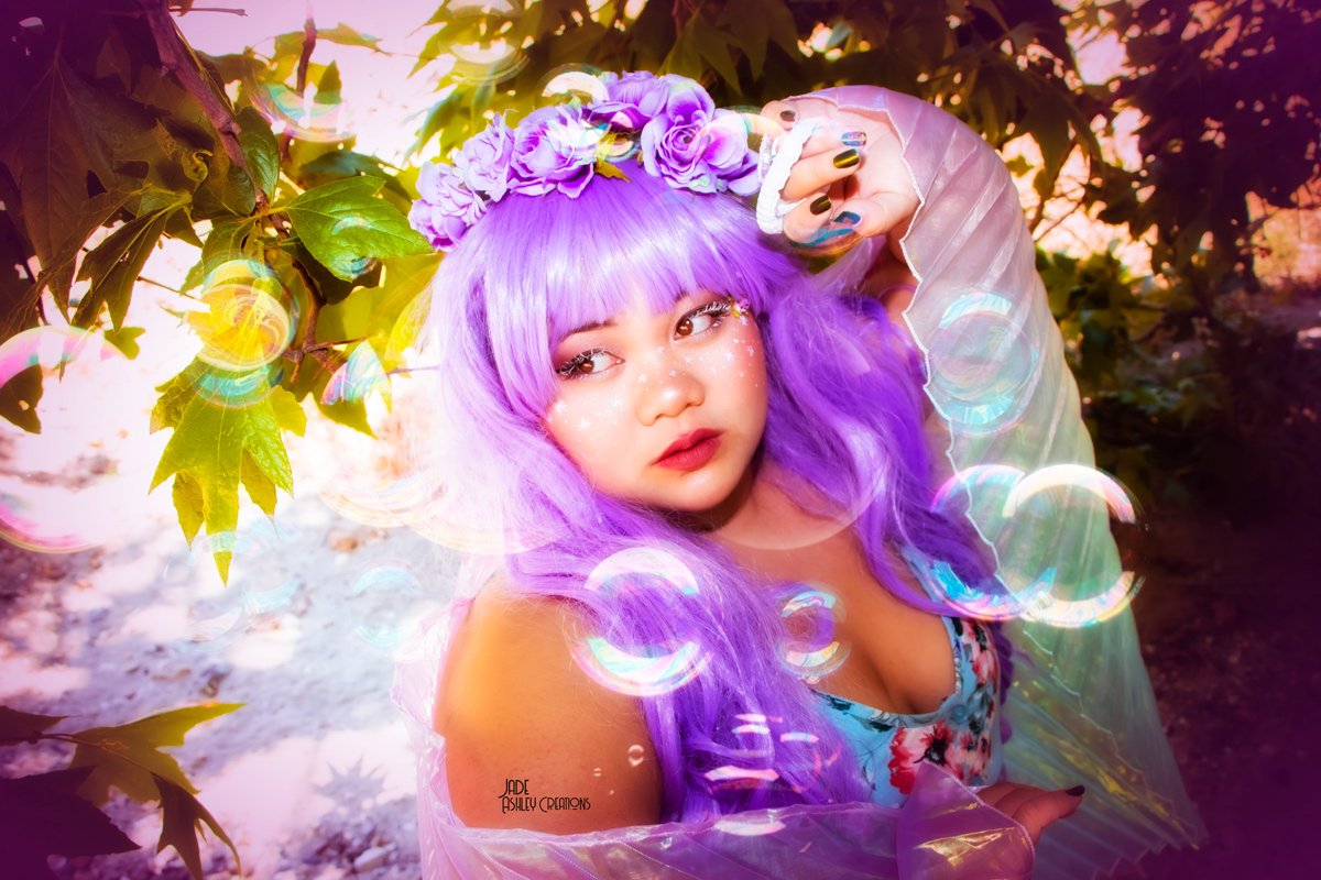 Young woman with purple hair, wearing a floral crown and glitter on her face, stands outdoors among green leaves and branches, with colorful bubbles in the foreground and background.