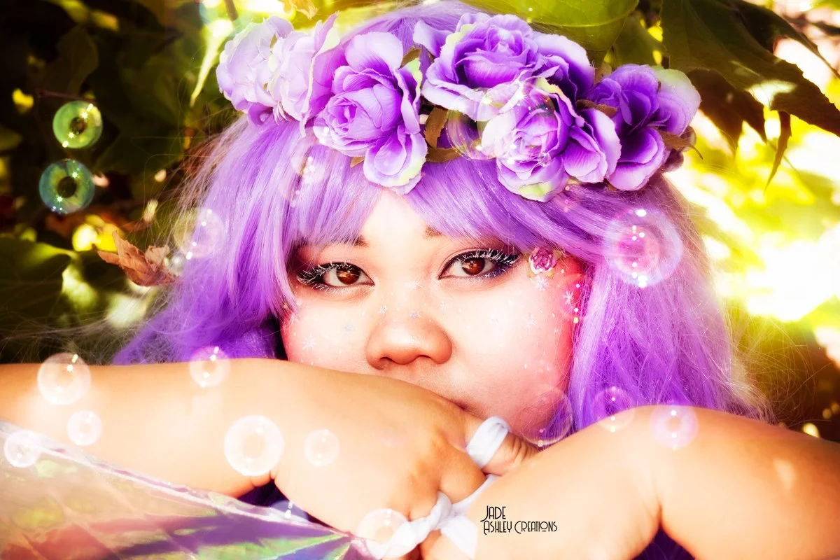 A person with purple hair and a purple flower crown looking at the camera, surrounded by greenery and glowing orbs.