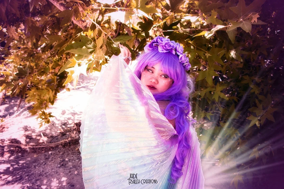 A woman with long, wavy purple hair, wearing a flower crown and glitter on her face, standing outdoors among trees with sunlight filtering through the leaves, holding a glowing iridescent fabric.