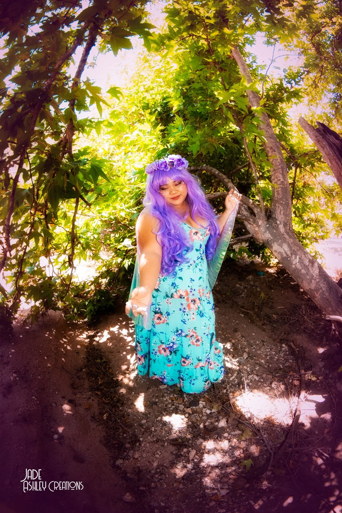 A woman with long purple hair and a purple flower crown wears a blue floral dress, standing outdoors among green trees with sunlight filtering through.