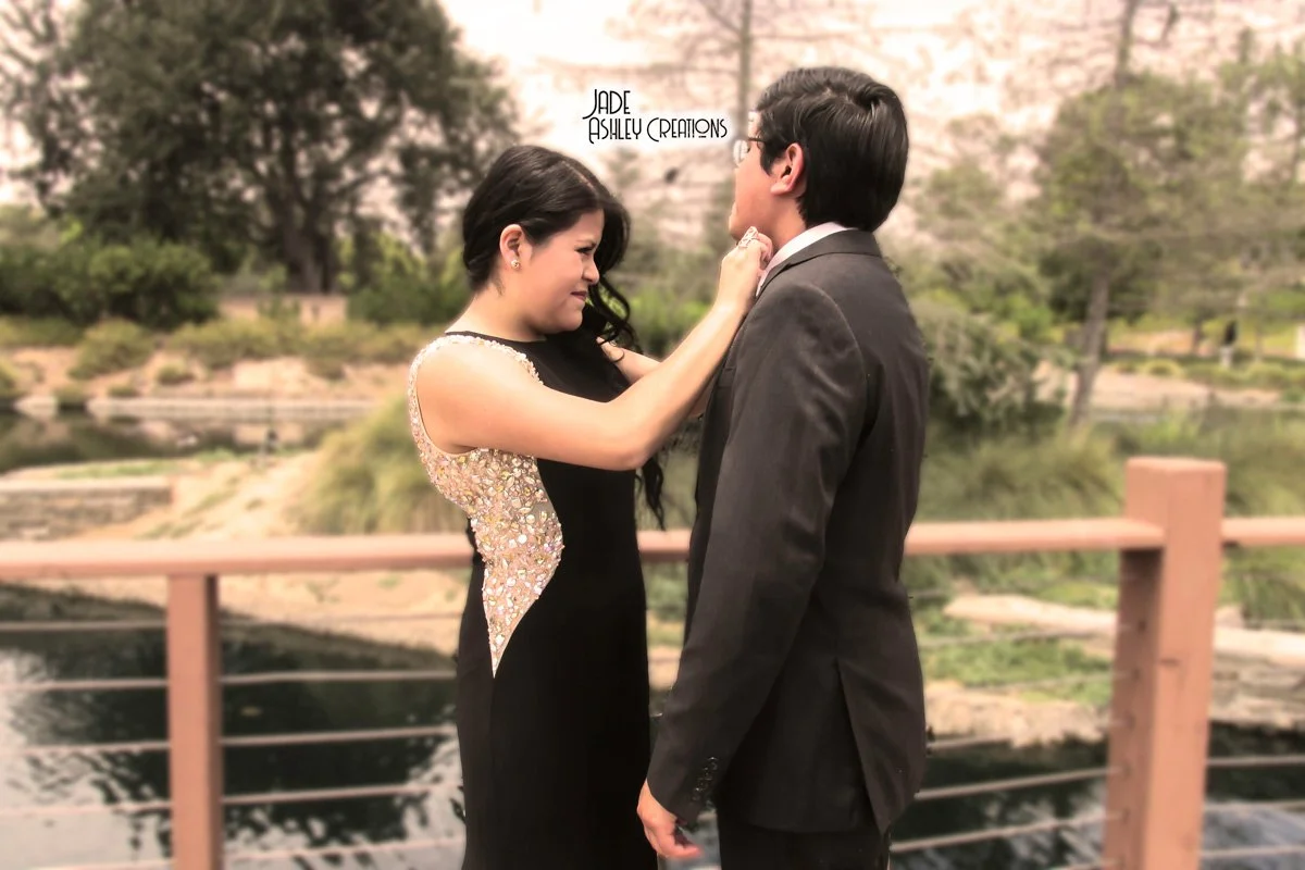 A woman and a man on a bridge, gazing into each other's eyes, with the woman touching the man's face. The woman has long black hair, wears a black dress with gold embellishments on the side, and has her eyes closed. The man has dark hair, glasses, an