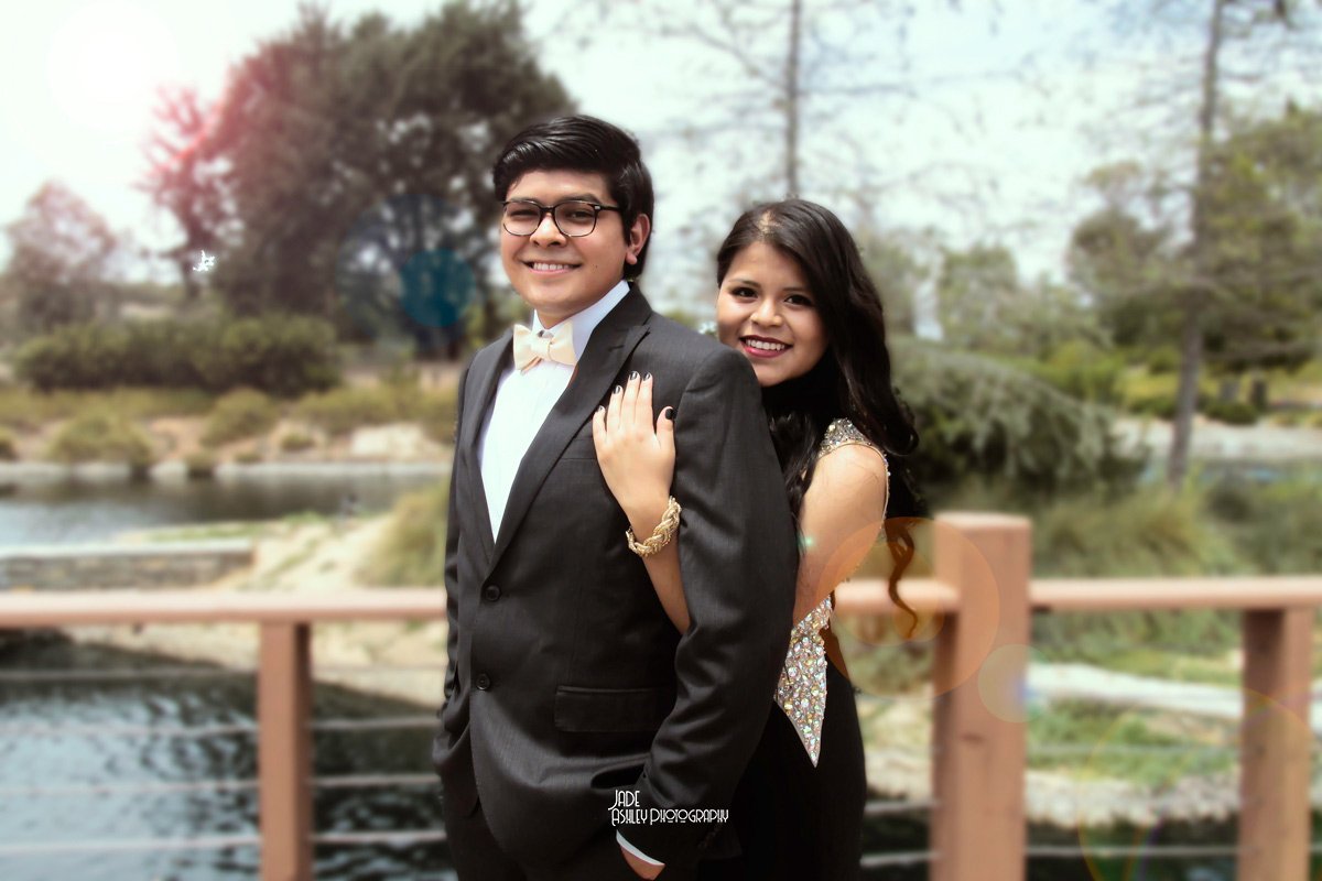 A young couple dressed in formal attire standing outdoors on a bridge with a nature scene in the background. The man is wearing a black suit with a white shirt and light-colored bow tie, and the woman is wearing a sleeveless dress with sparkly detail