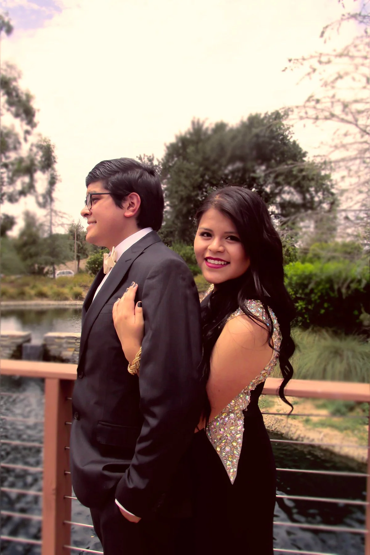 A young man in a tuxedo and a woman in a sparkly dress are standing outdoors near a body of water and greenery, smiling at the camera.